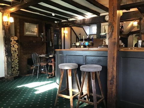 Interior view of a cozy traditional English pub with wooden furnishings, dim lighting, and a warm atmosphere. The bar is adorned with beer taps, glasses, and bottles, and there are bar stools and high tables with stools for customers to sit and enjoy their drinks.