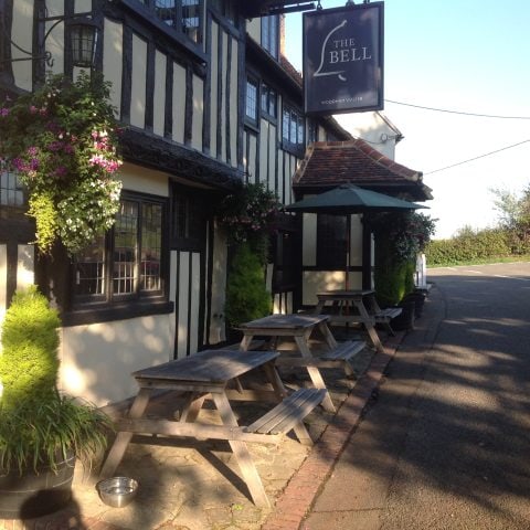 Exterior view of the Bell Pub, a charming English pub. Tables and chairs are set up outside the entrance, inviting customers to enjoy a pint in the sunshine.